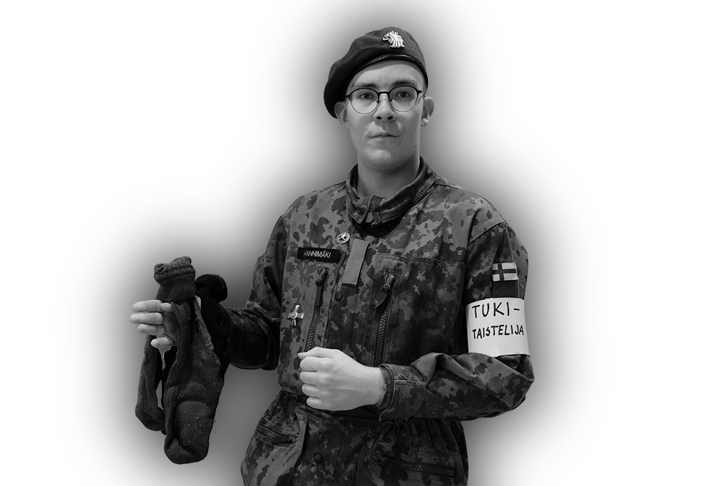 Kaartinjääkäri Hanhimäki katsoo kameraan ja pitää kädessään kahta sukkaparia. Toisessa kädessä hänellä on Tukitaistelija-tunnus.