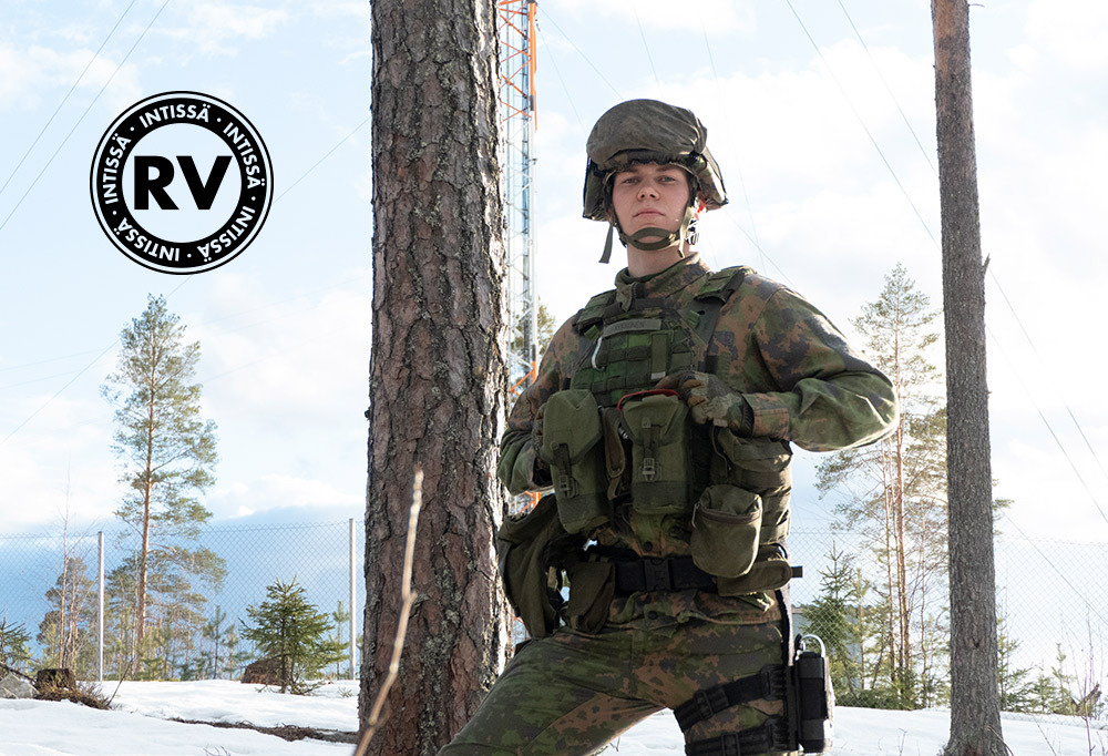 Varusmies seisoo puun edessä taisteluvarustus päällään.