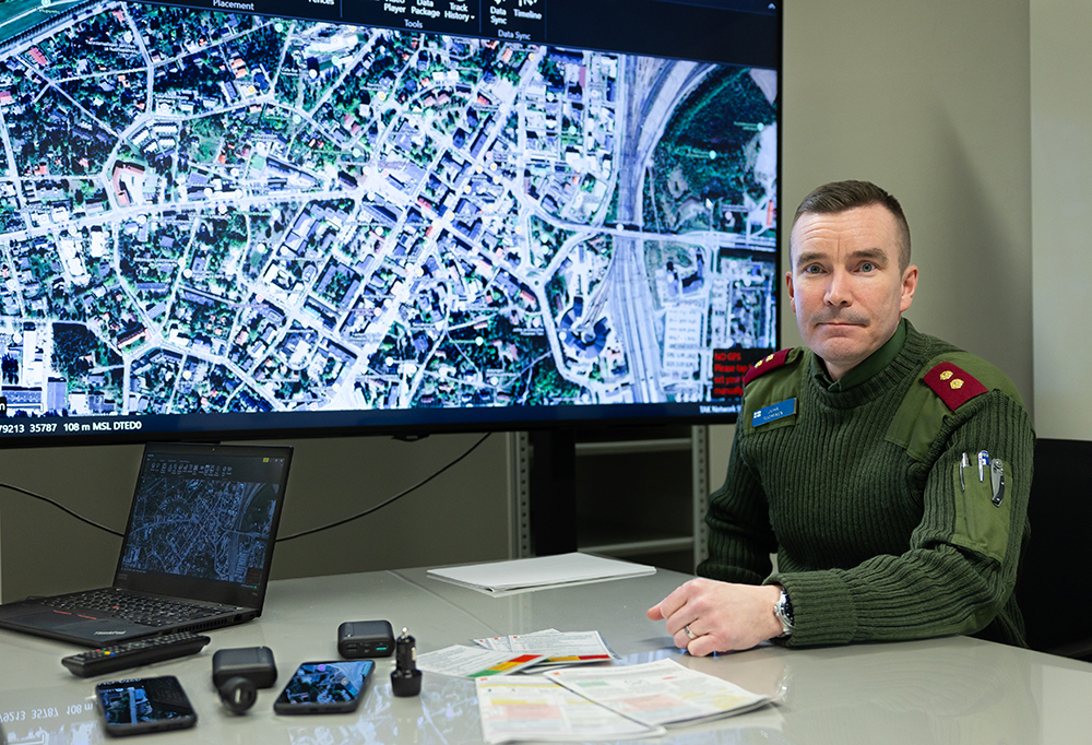 Everstiluutnantti Juha Tuominen katsoo kameraan. Taustalla näyttö, jossa esillä satelliittikuva kaupungista.
