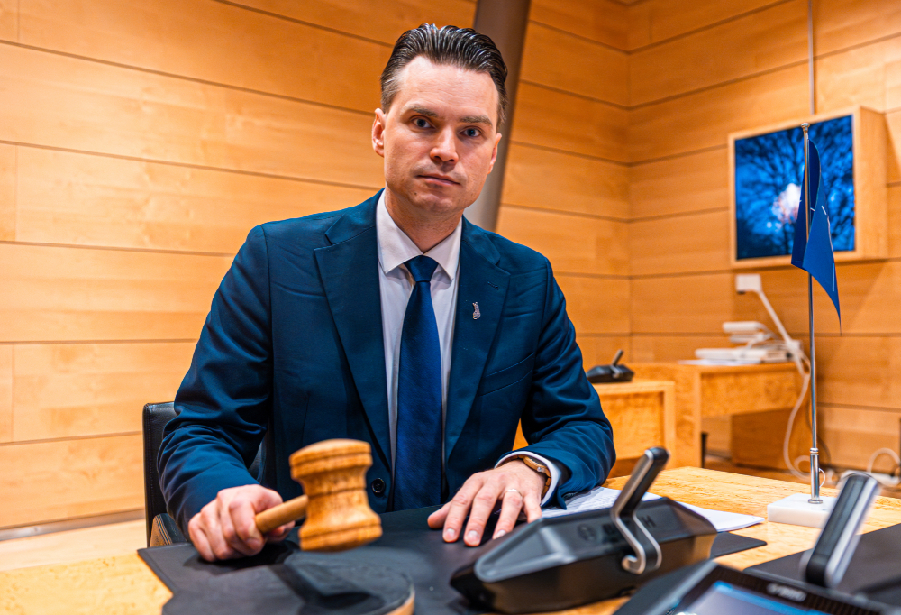 Heikki Autto katsoo kameraan istuen pöydän ääressä. Kädessä hänellä on puheenjohtajan nuija ja pöydällä Nato-lippu.