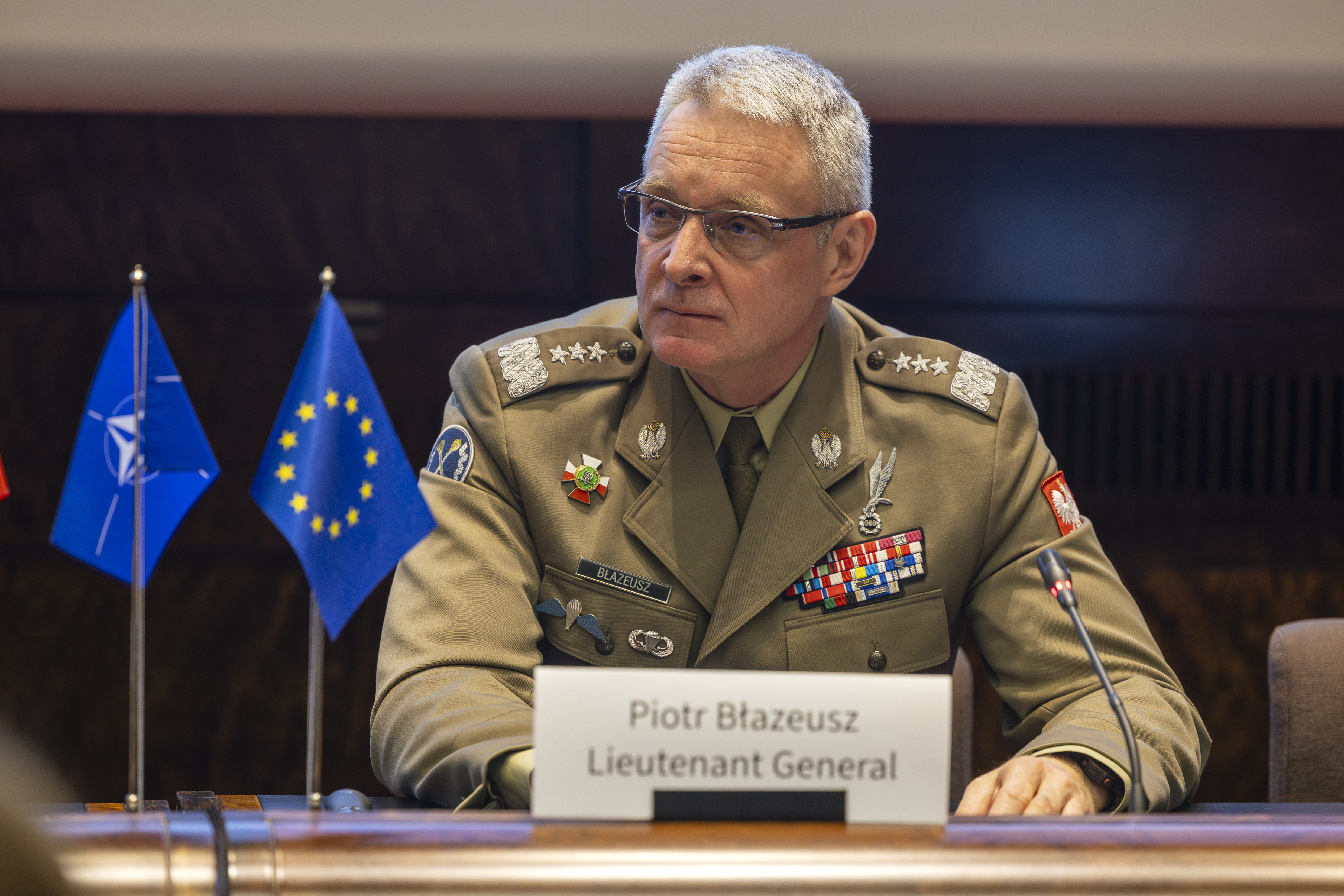 Kuvassa puolalainen kenraaliluutnantti Piotr Blazeusz istuu mikrofonin edessä ja katsoo kuvan vasempaan nurkkaan. Pöydällä on Naton ja EU:n liput.