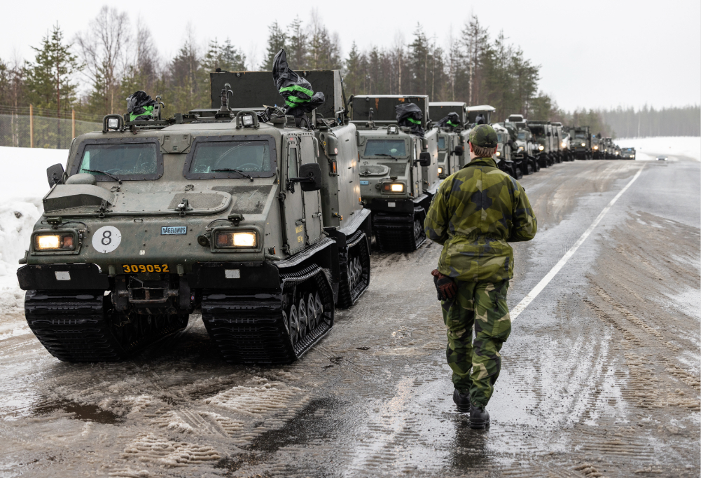 Kuvassa on jonossa useampi panssarivaunu. Tiellä kävelee Puolustuvoimien vihreään asuun pukeutunut henkilö.