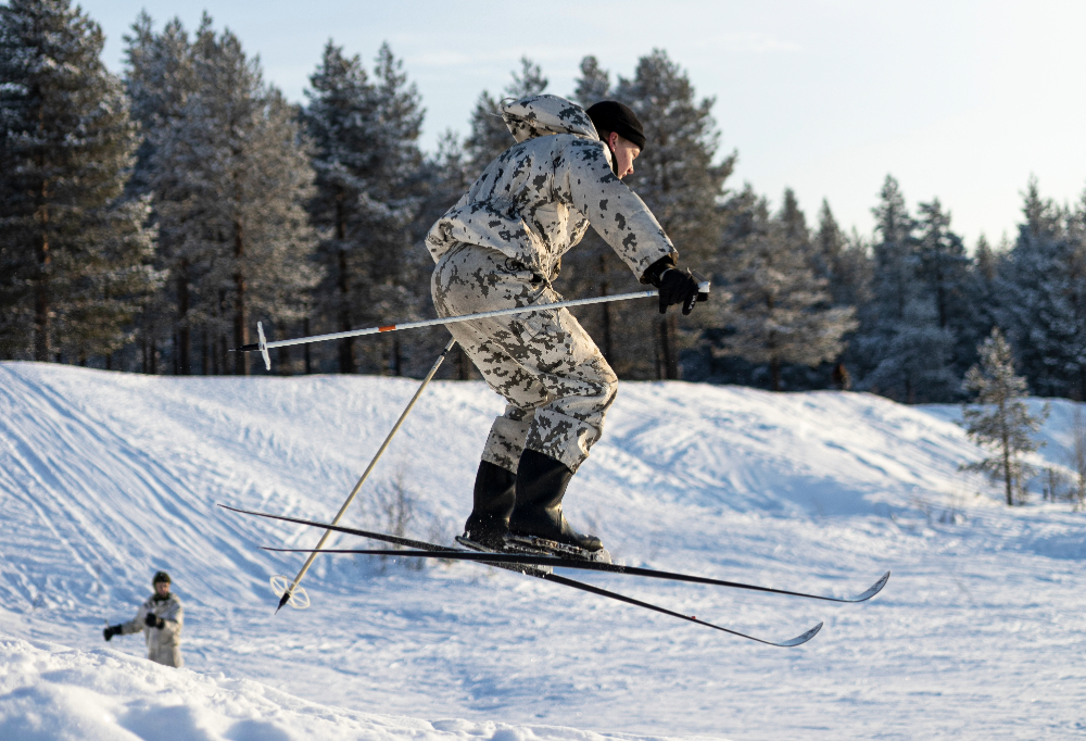 Lumipukuun pukeutunut taistelija hyppää suksiensa kanssa. Taustalla luminen metsä.