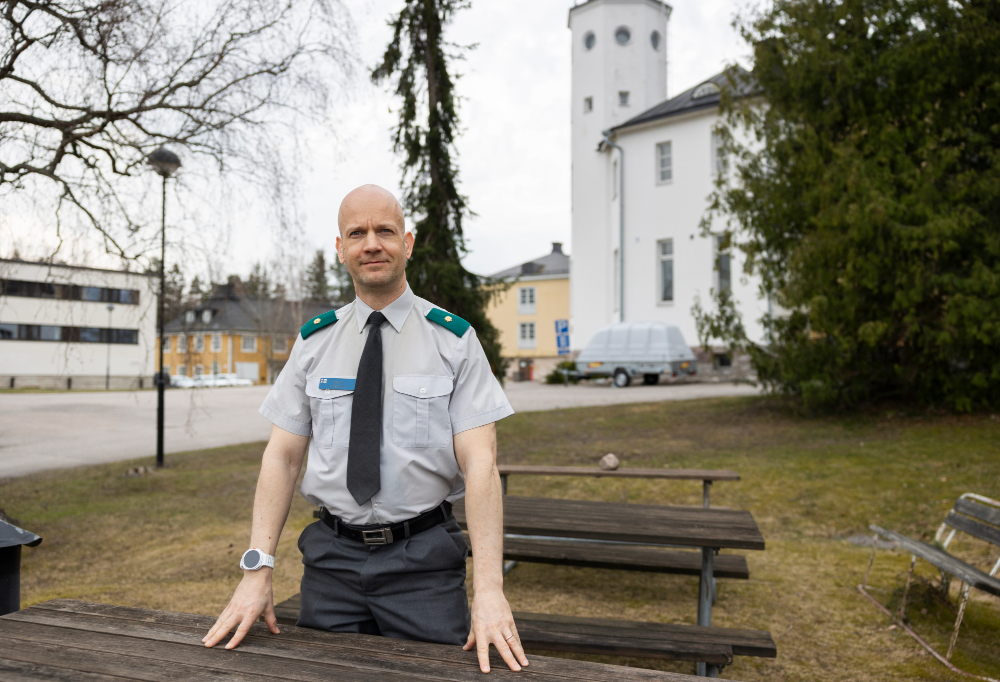 Majuri Kostian seisoo pöytä-penkin ääressä ja katsoo kameraan. Taustalla vaaleita, kaksikerroksisia rakennuksia.