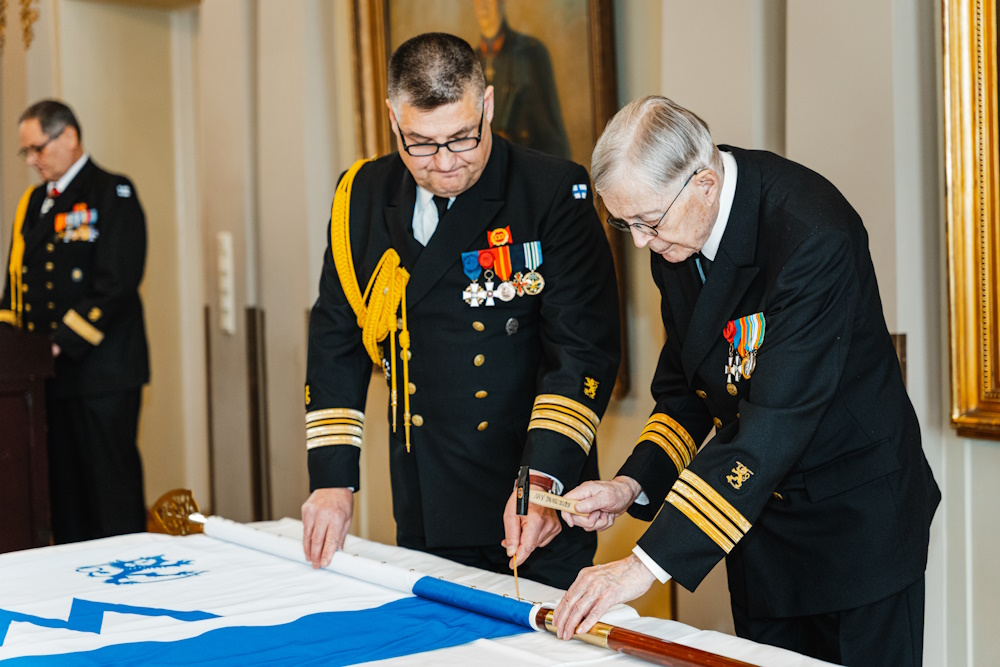 Ruotuväen entinen päätoimittaja komentajakapteeni evp Tapani Talari lyö naulansa lipputankoon. Komentaja evp Mikko Saarinen osoittaa naulan mihin lyödä.