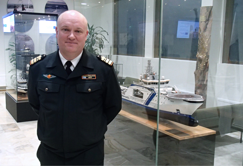 Komentaja Mikko Hirvi seisoo kädet selkänsä takana ja katsoo kameraan. Taustalla pienoismalleja Rajavartiolaitoksen aluksista.