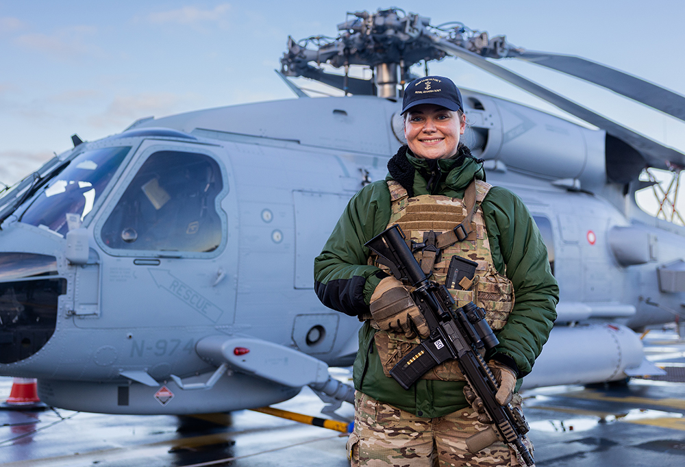 Taistelija hymyilee kameralle kivääri käsissään. Taustalla taisteluhelikopteri.