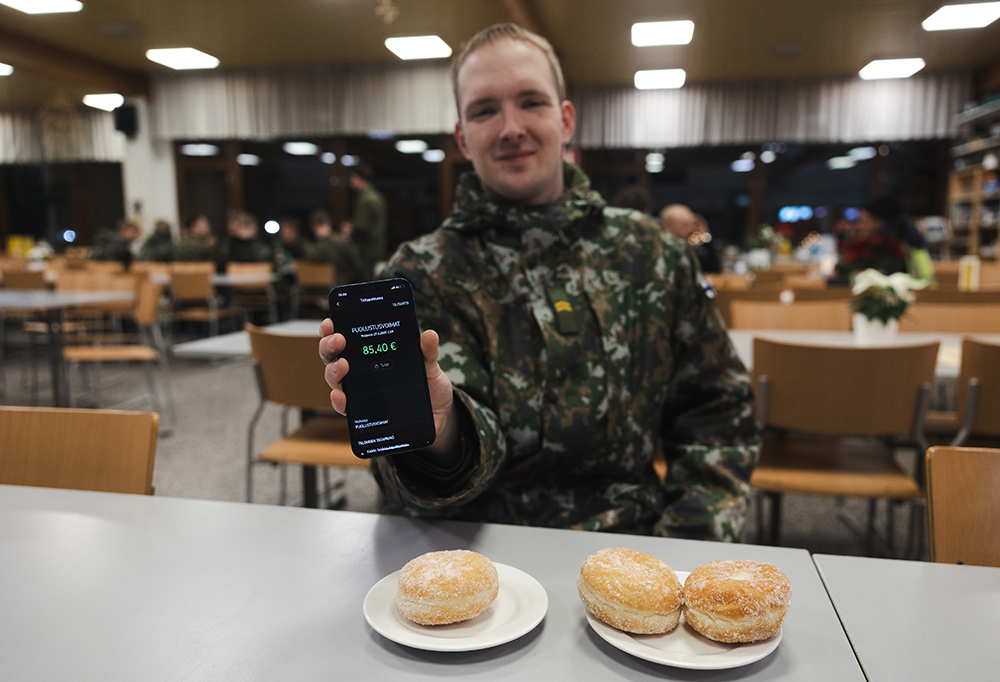Alikersantti Marjamäki näyttää kameralle Puolustusvoimilta tullutta maksusuoritetta. Pöydällä kolme munkkia.