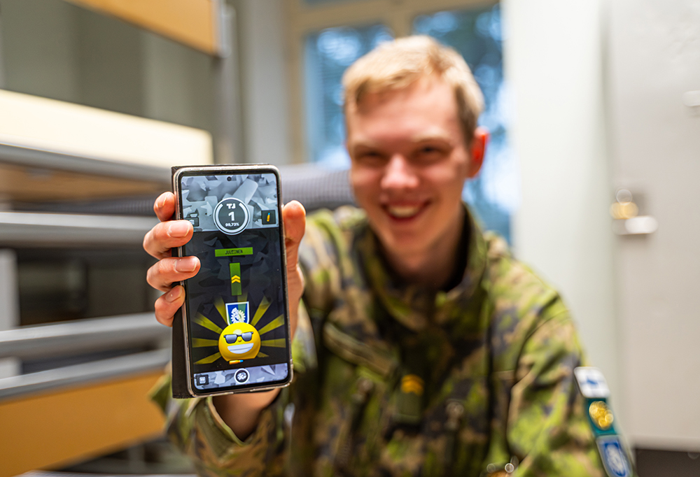 Alikersantti Juutinen näyttää kameralle kännykkää kasarmin tuvassa. Näytöllä näkyy Intti-sovellus, jossa näkyy hymyilevä 