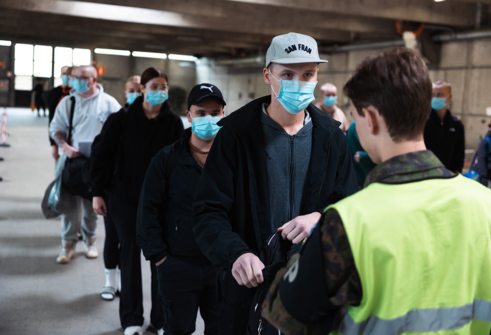 Tulevia varusmiehiä jonottaa sisätiloissa kasvomaskit päällä. Sotilaspoliisi tarkastaa ensimmäisen jonottajan repun.