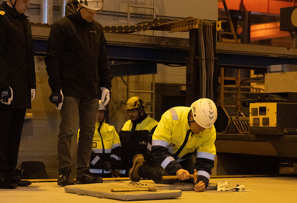 Kuvassa telakan työntekijä stanssaamassa polvillaan teräslevyn päällä. Taustalla kutsuvieraita ja telakkahallia.