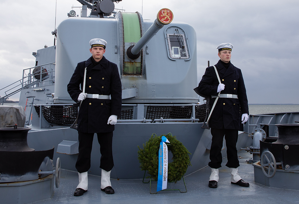 Kaksi taistelijaa seisoo seppeleen molemmin puolin miinalaivan kannella.