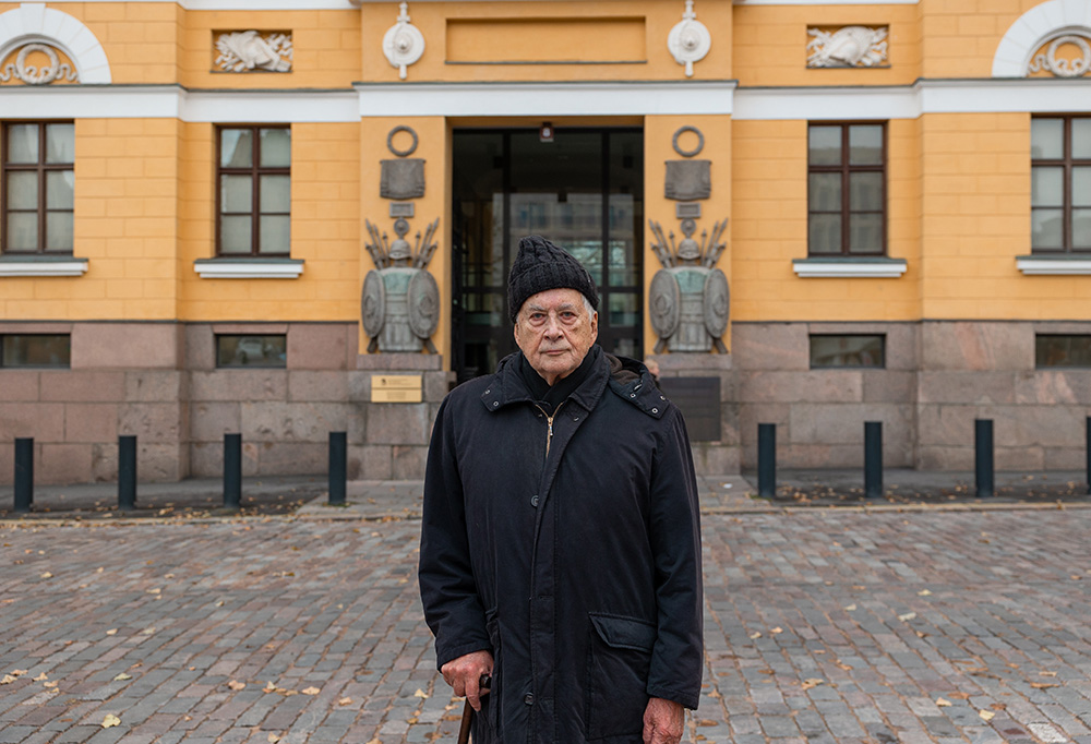 Gustav Hägglund katsoo vakavana kameraan. Hän seisoo keltaisen tiilitalon edessä.