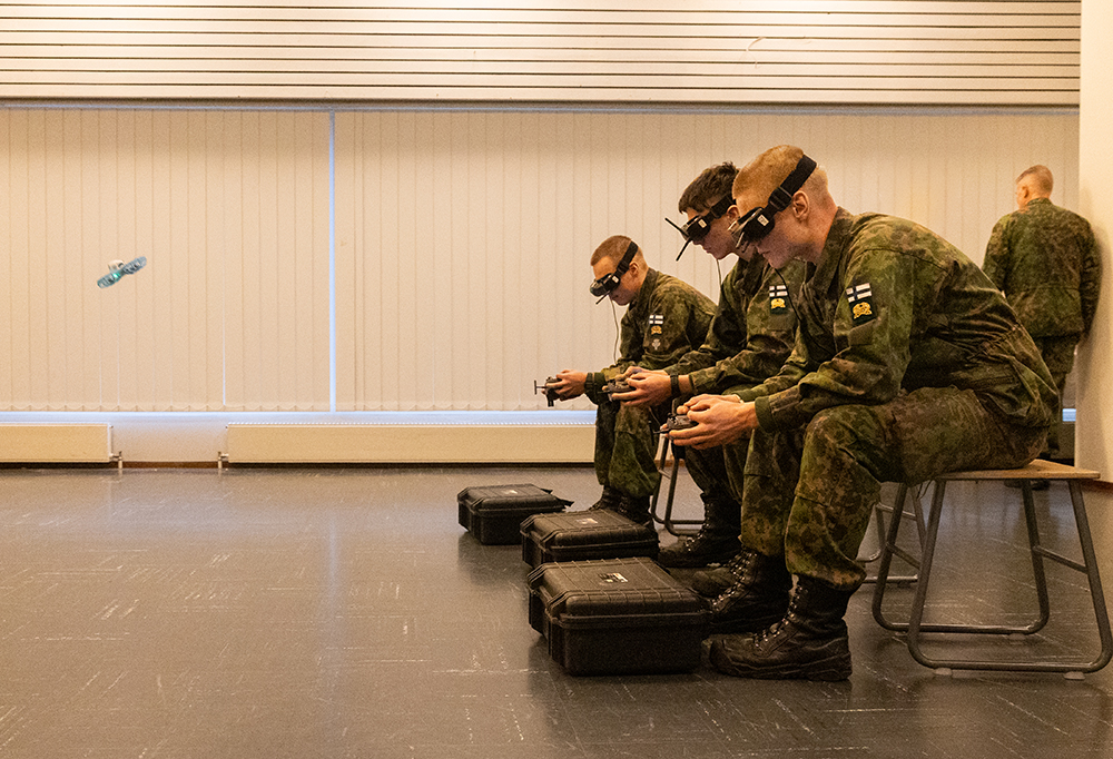 Varusmiehiä istuu rivissä taistelujakkaroilla keskellä suurta huonetta, ja ohjaavat FPV-mikrolennokkeja. Yksi mikrolennokki näkyy kuvassa.