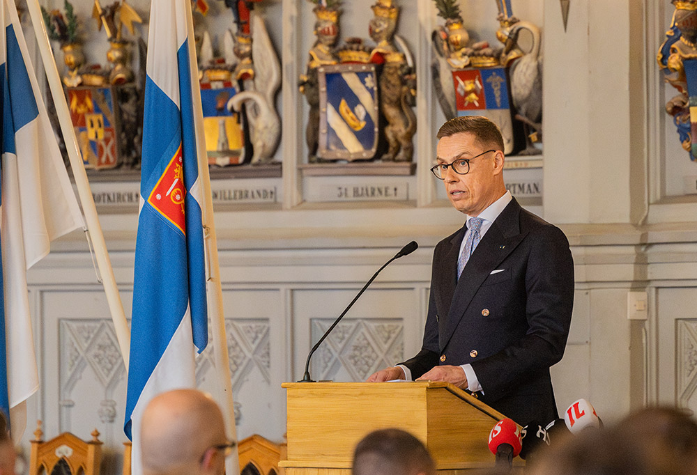 Alexander Stubb puhumassa mikrofoniin puhujanpöntössä. Etualalla yleisöä, vasemmalla kaksi lippua ja taustalla vaakunoita.