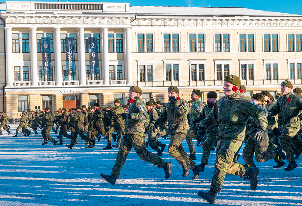 Upseerikokelaat juoksevat ruusut suissaan kohti seuralaisiaan, keltaisen, koristeellisen rakennuksen ohi.