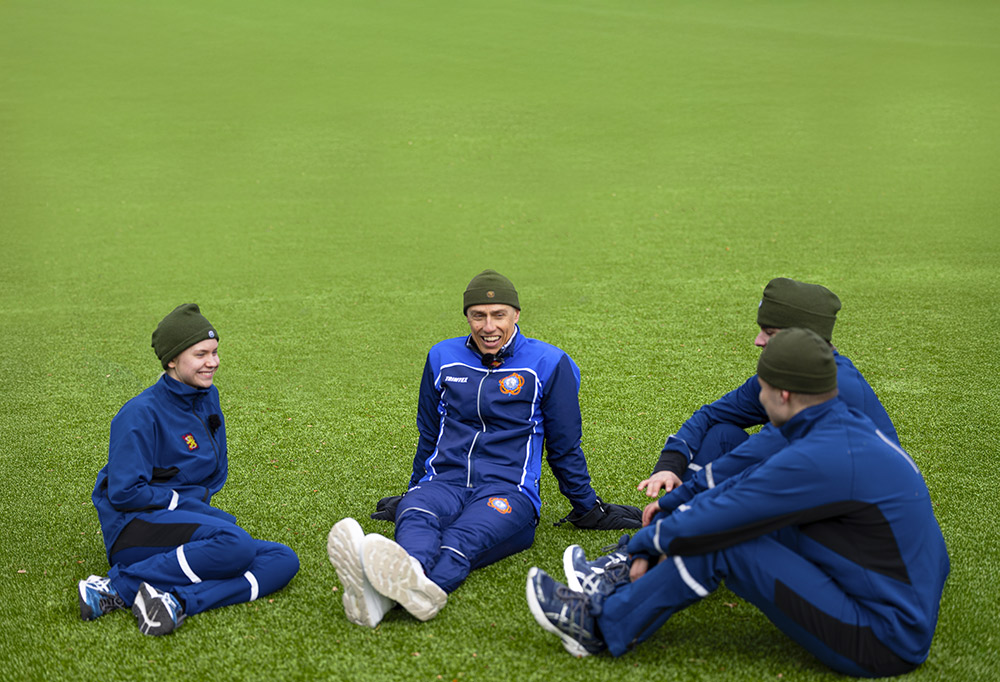 Tasavallan presidentti Alexander Stubb istuu ja hymyilee tekonurmikentällä kolmen varusmiespalvelusta suorittavan ympäröimänä.