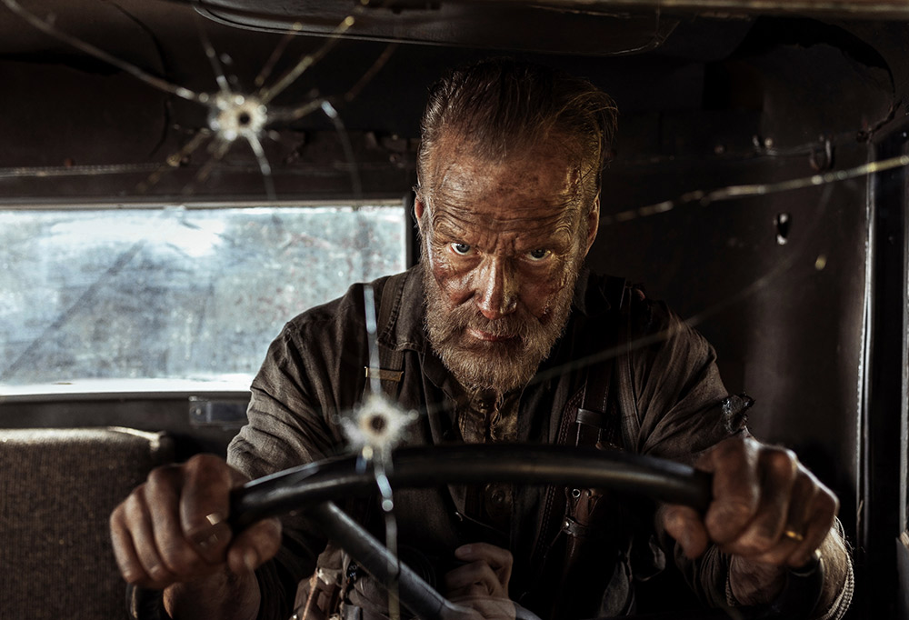 Parrakas mies ajaa autoa. Auton tuulilasissa on ammuksien aiheuttamia reikiä.