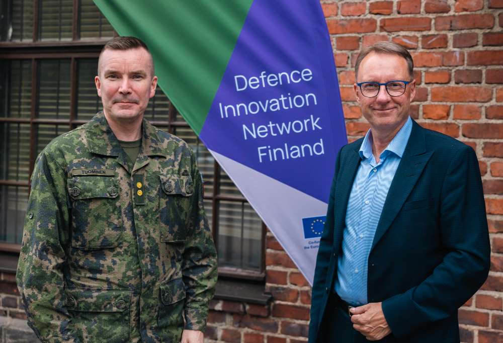 Everstiluutnantti ja kenraaliluutnantti poseeraavat tiiliseinän edessä. Taustalla Definen mainosbanneri.