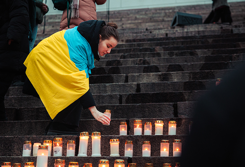 Ukrainan lippua selässään pitävä nainen laskee kynttilää kiviportaille.