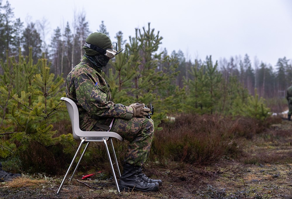 Kuvassa varusmies istuu metsässä tuolilla drone-ohjain kädessään.