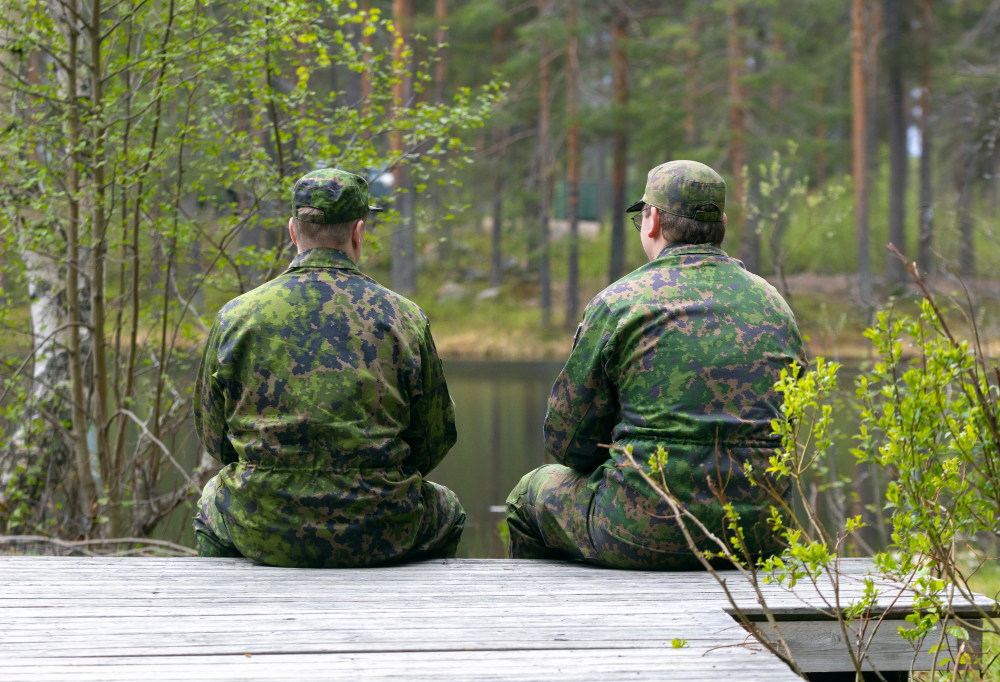 Kaksi sotilasta istuvat selin kameraan toisiaan kohti kääntyneinä luontomaisemassa.