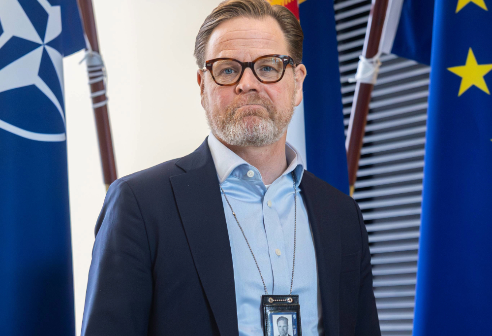 Kuvassa Puolustusministeriön kansliapäällikkö Janne Kuusela seisoo lippujen edessä ja katsoo kameraan puku päällä.