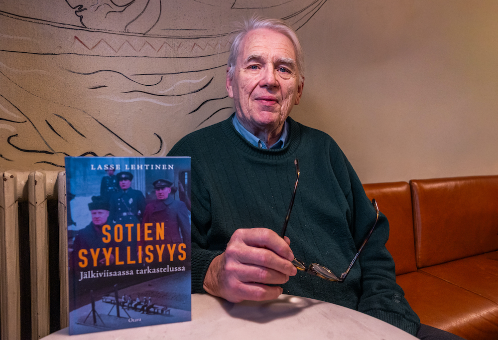 Lasse Lehtinen katsoo kameraa Sotien syyllisyys -kirja vierellään.