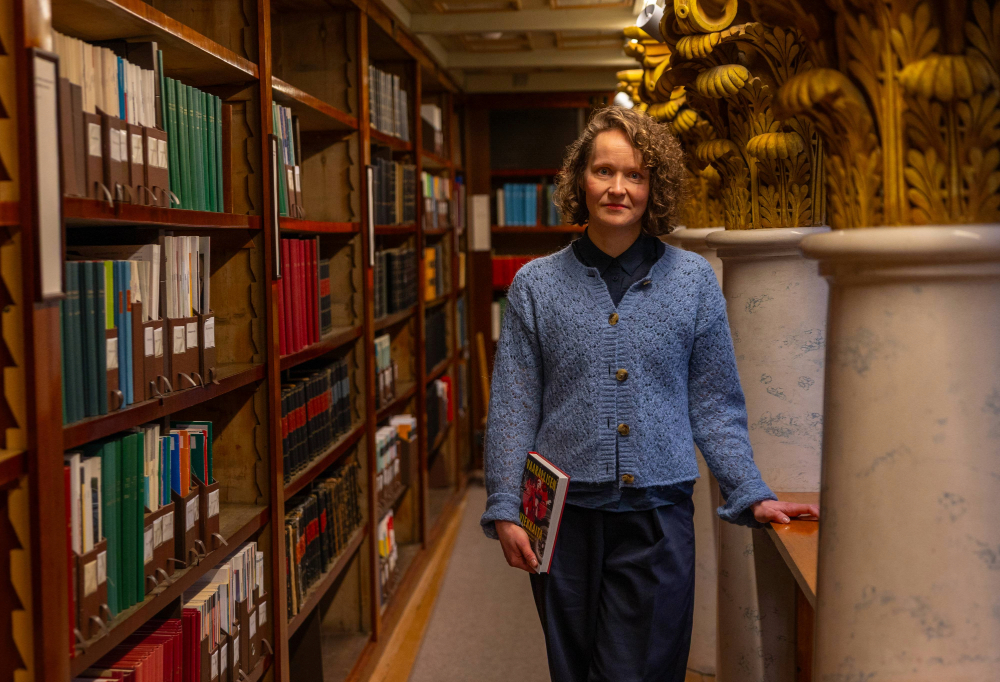 Kuvassa Turun yliopiston poliittisen historian tutkija Piia Vuorinen seisoo kirja kädessään katsoen kameraan ja nojaten puutasoon.