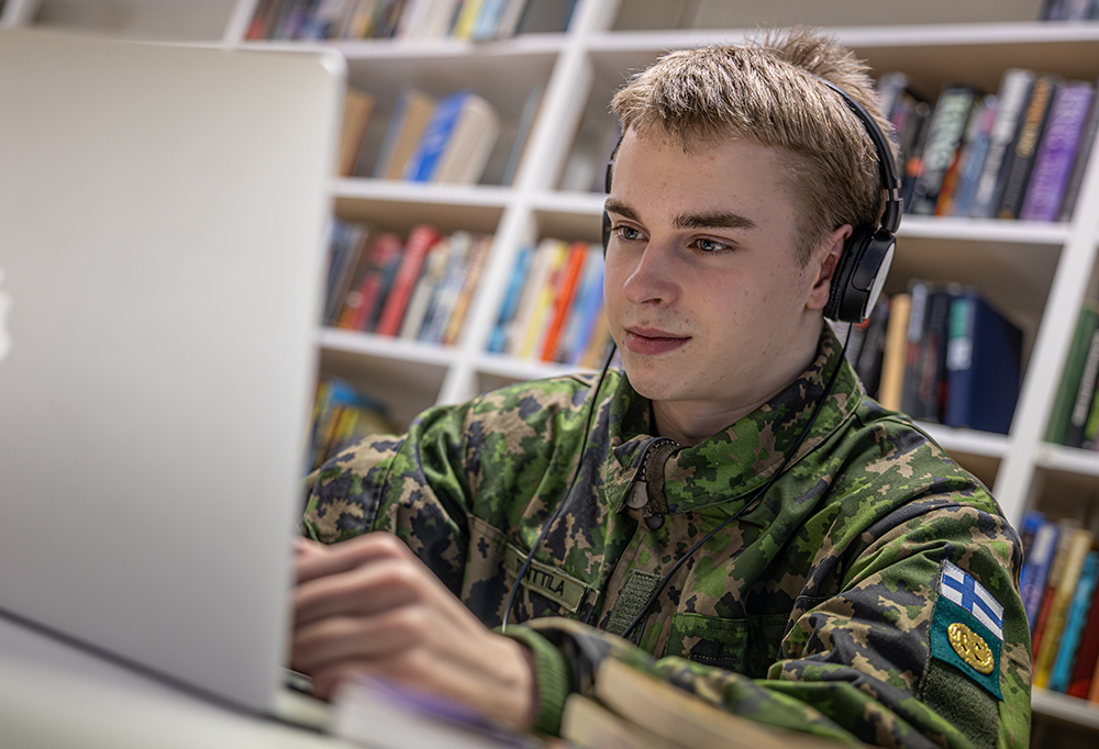 Kuvassa varusmies katsoo keskittyneesti tietokoneen ruutua kuulokkeet päässä.