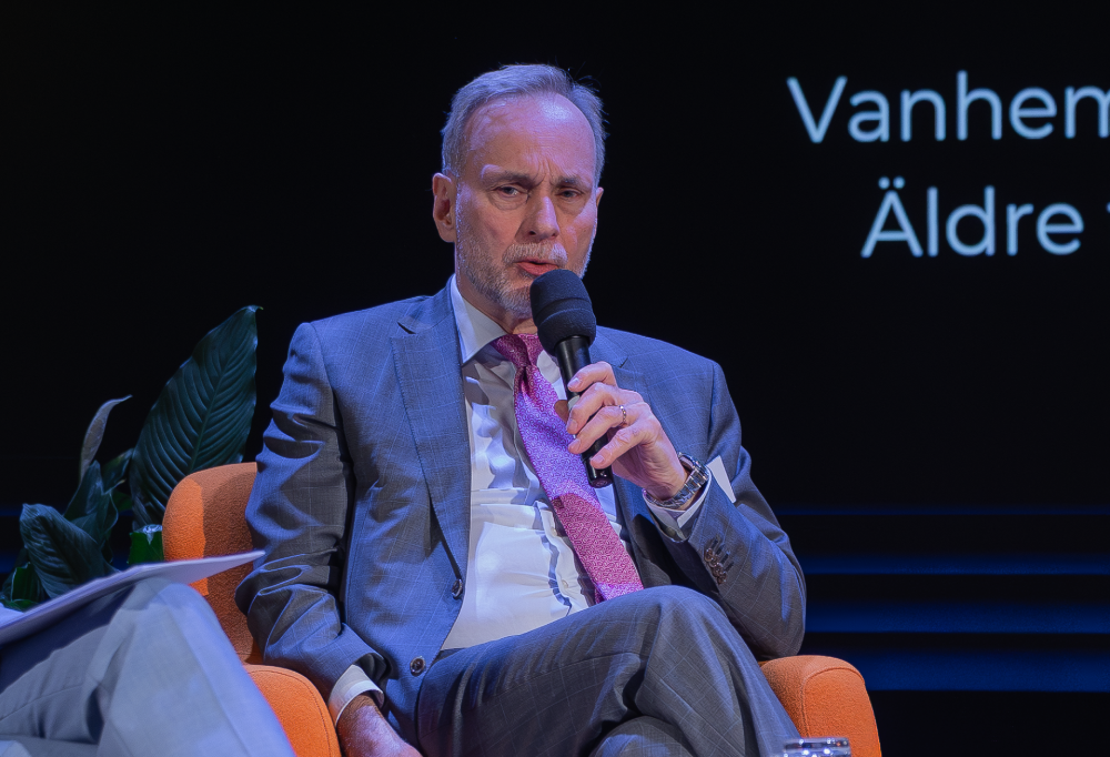 Rautalinko puhumassa paneelissa mikrofoniin. Hän istuu oranssilla nojatuolilla.