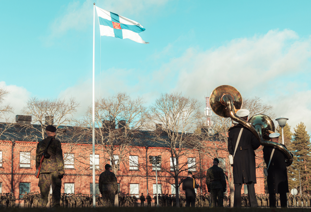 Puolustusvoimien juhlatilaisuudessa lippu liehuu ja pihalla on maastokuvioon pukeutuneita ihmisiä.