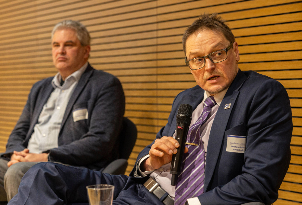 Juha-Matti Lehtonen puhuu mikkiin. Taustalla toinen puhuja ja puinen seinä.