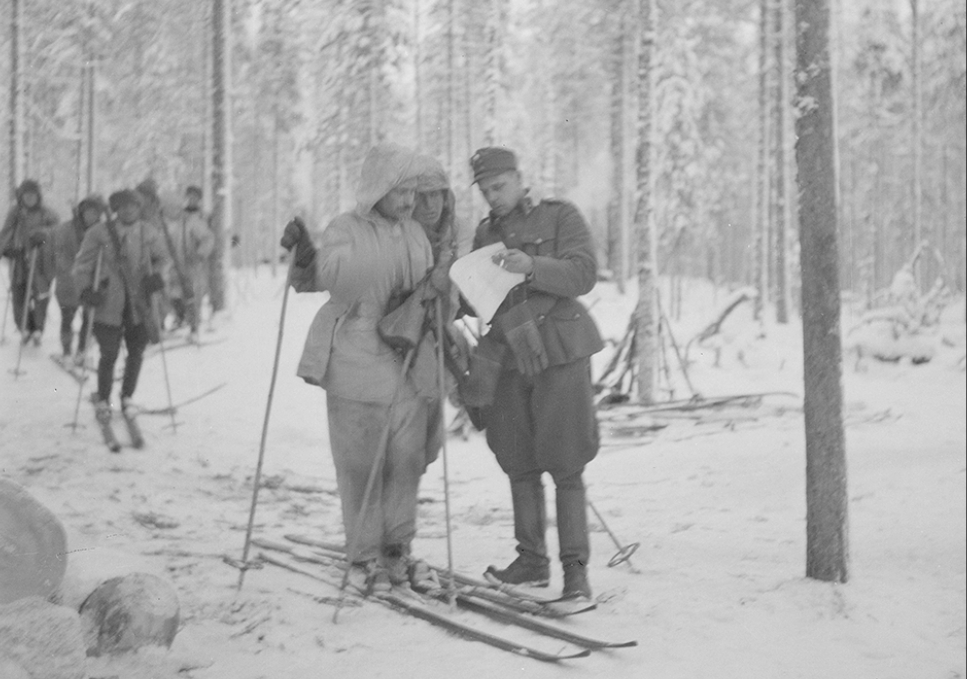 Taistelijoita hiihtämässä talvisessa metsässä. Yhdellä on kädessään kartta, jota kaksi muuta taistelijaa lukevat.
