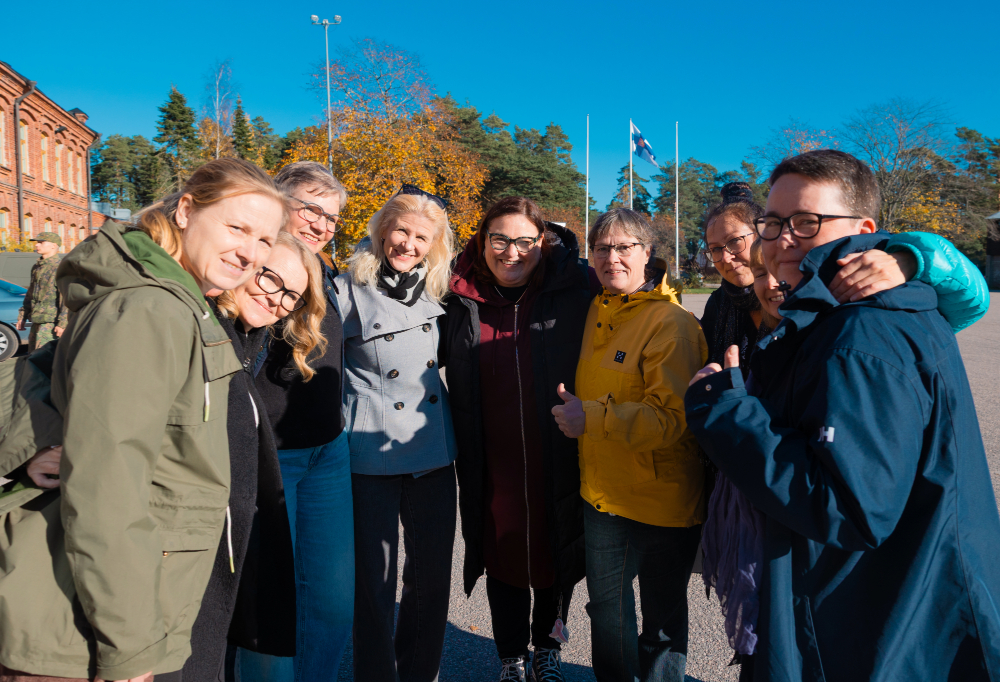 Naiset halaavat puolikaaressa pihalla. Taivas on sininen ja taustalla heiluu Suomen lippu.