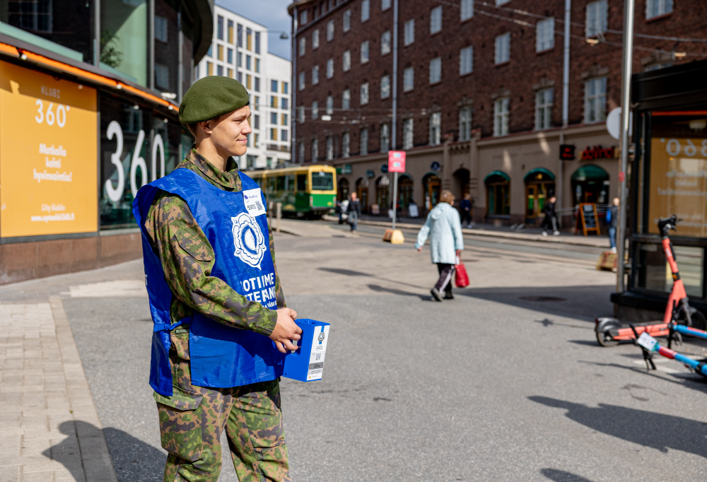 Varusmies seisoo kädessään varainkeruulipas. Ympärillä näkyy keskustaympäristöä.