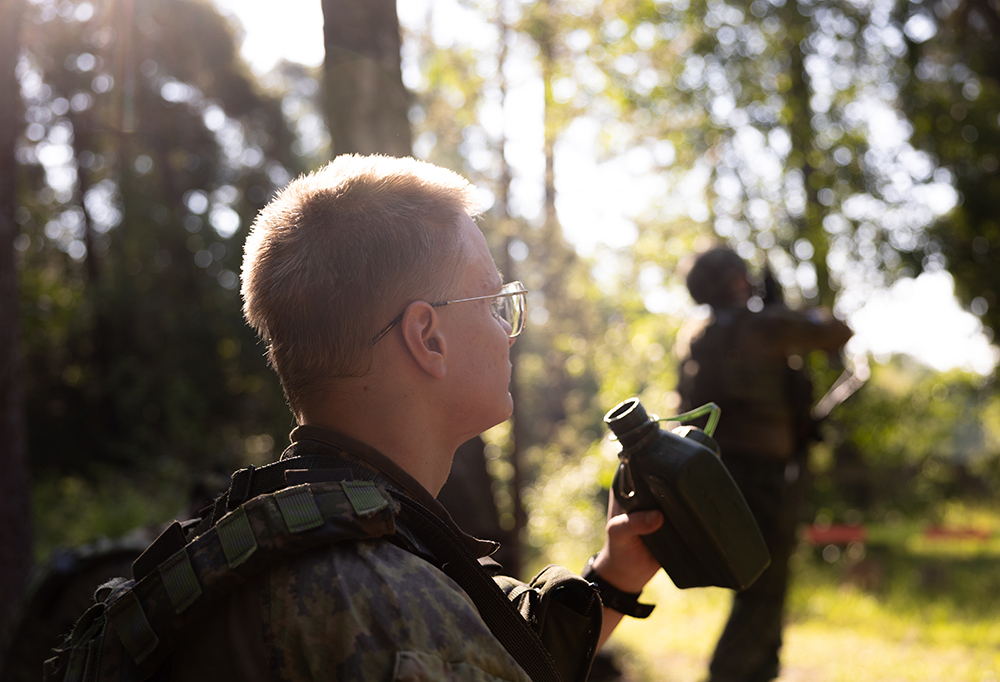 Varusmies istuu varjoisalla taukopaikalla ilman kypärää. Hän on nostamassa kenttäpulloa huulilleen.