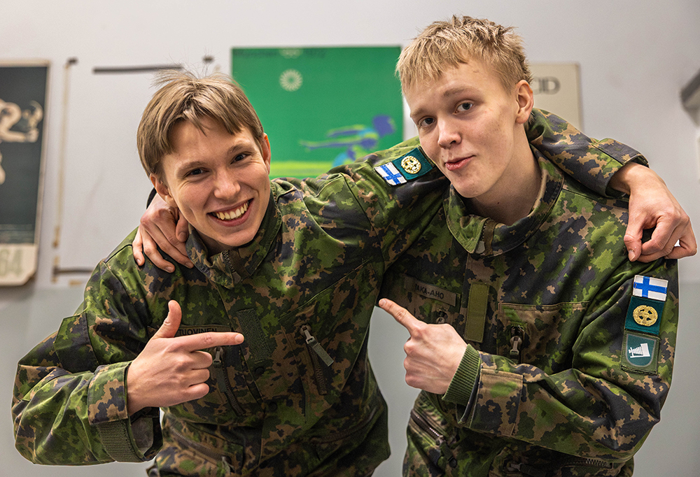 Kuvassa kaksi varusmiestä osoittavat toisiaan sormella ja hymyilevät.