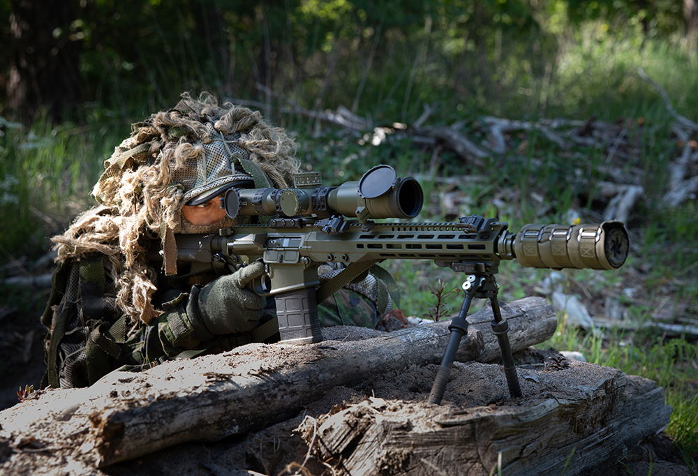 Kuvassa metsään maastoutunut mies tähtää asellaan poterosta.