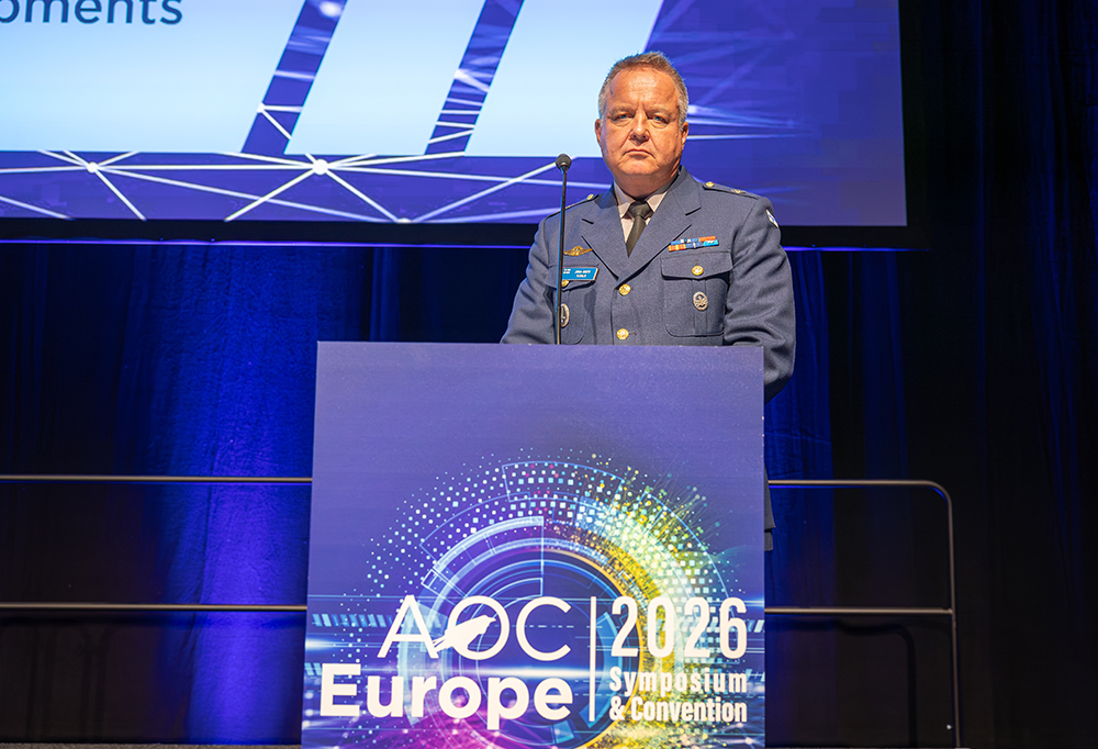Kuvassa insinööriprikaatikenraali Juha-Matti Ylitalo katsoo kameraan puku päällä AOC Europe-puhujankopin takaa.