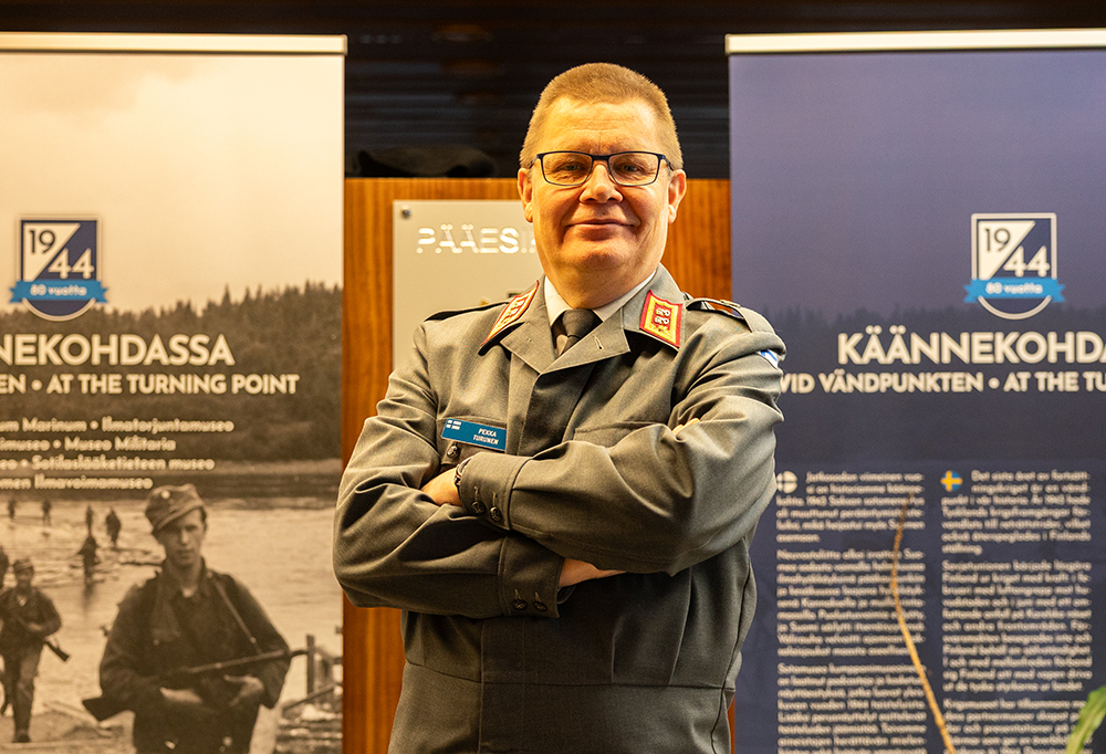 Kenraalimajuri Pekka Turunen seisoo kahden bannerin edessä ja katsoo kameraan kädet puuskassa.