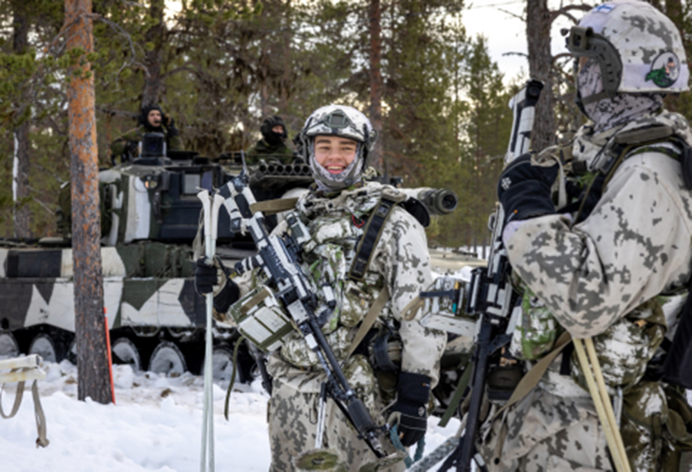 Lumipukuun pukeutunut sotilas hymyilee talvisessa metsässä taistelukaverilleen. Taustalla panssarivaunu.